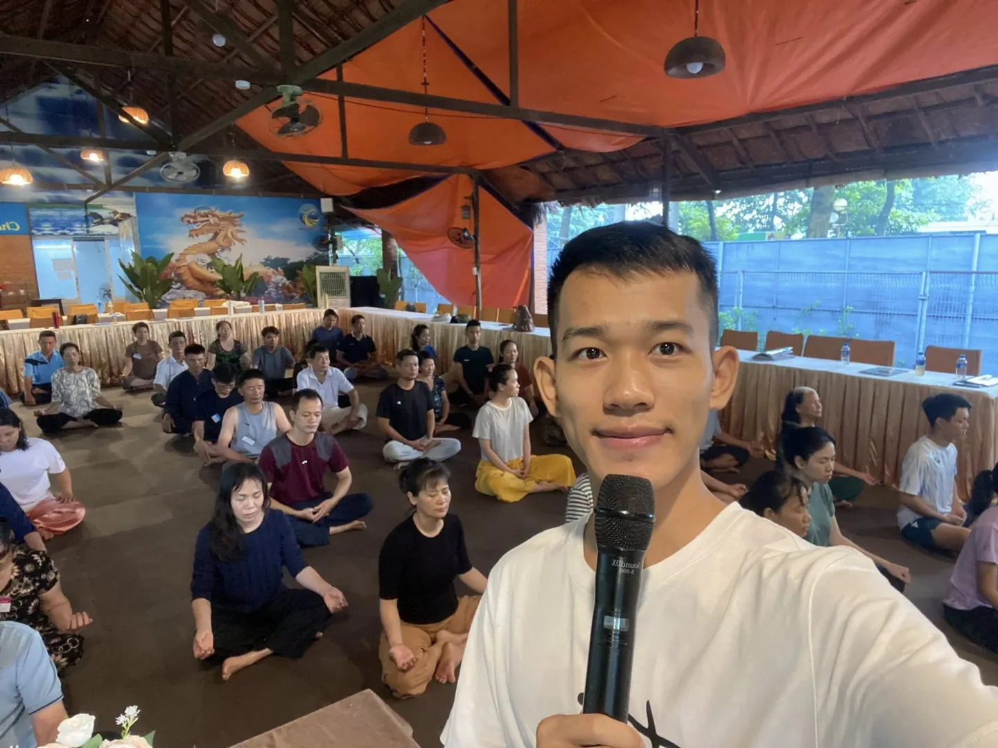 Tâm Đức An hướng dẫn thở thiền cho học viên ngồi kiết già trong một hội trường mái lá