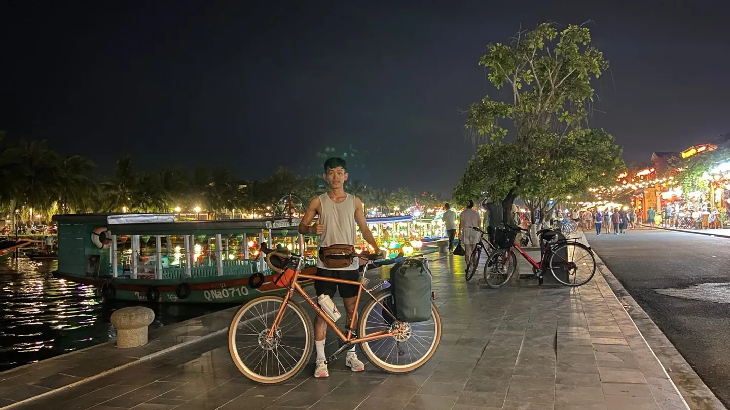 Tâm Đức An đứng cạnh chiếc xe đạp có túi hành lý bên bờ sông Hoài, Hội An về đêm